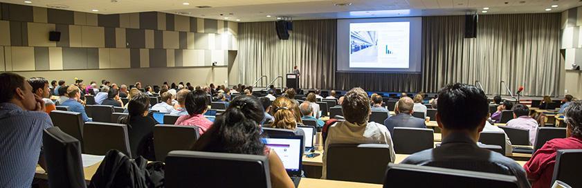 A room filled with people gazes toward a stage, where a speaker stands at a podium beside a large screen.